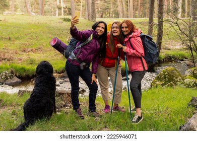 Three Women With Hiking Backpacks And A Dog Taking A Selfie While Trekking In A Forest With A River