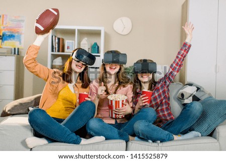 Three women in modern virtual reality headsets having expirience in watching football match at home. Girls in vr glasses sitting on the sofa and eating popcorn