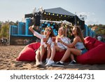 Three women sit on red bean bags, sunlit beach as backdrop, holding drinks, enjoying music festival. Smiling ladies in sunglasses relax by stage, casual summer outfits, friends at seaside.