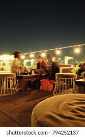 Three Women Sitting On A Chic Rooftop Bar At Night In Manhattan. Stools And Table, Large String Lights Along The Balcony, Lights Of New York City In The Background. Low Light Photography, Motion Blur.