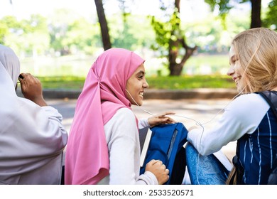 Three women sitting outdoors, sharing earphones. Two women in hijabs, one in a pink hijab, smiling. Diverse group enjoying music in a park setting. Young Muslim woman in pink hijab with friends. - Powered by Shutterstock