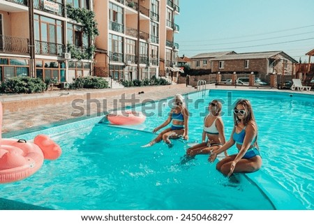 Three women are sitting in a pool with pink and blue floaties. Scene is lighthearted and fun.