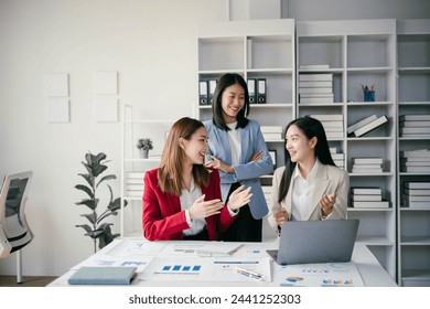 Three women are smiling and talking in a business setting. They are dressed in business attire and are standing in front of a white desk with a laptop. The room is filled with bookshelves - Powered by Shutterstock