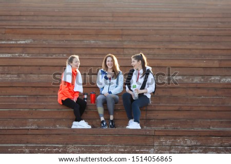 Three women in sportswear after a workout sitting on wooden steps in a summer city park. Sunset. Back light.