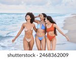 Three women are walking on the beach, smiling and enjoying their time together. The beach is calm and the water is a beautiful blue color. The women are wearing colorful swimsuits