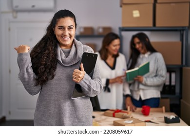 Three Women Working At Small Business Ecommerce Pointing Thumb Up To The Side Smiling Happy With Open Mouth 