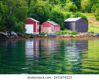 three wooden boat huts on the shore of a Norwegian fjord with reflections in the water - Powered by Shutterstock