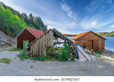 Three wooden structures, two intact sheds and one in disrepair, sit on a serene coastal landscape. Lush greenery and tranquil waters surround the scene under a beautiful clear blue sky.