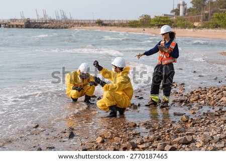 Three workers in bright rain gear and hard hats check water samples at the edge of the sea. They are on the shore, with a pier and cranes in the background. It is daytime and sunny.