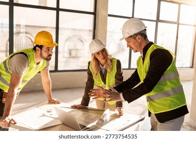 Three workers gather around a table to discuss project plans. They look at blueprints and a laptop, exchanging ideas in a bright construction site.