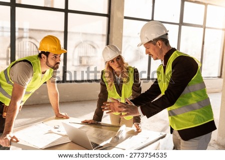 Three workers gather around a table to discuss project plans. They look at blueprints and a laptop, exchanging ideas in a bright construction site.