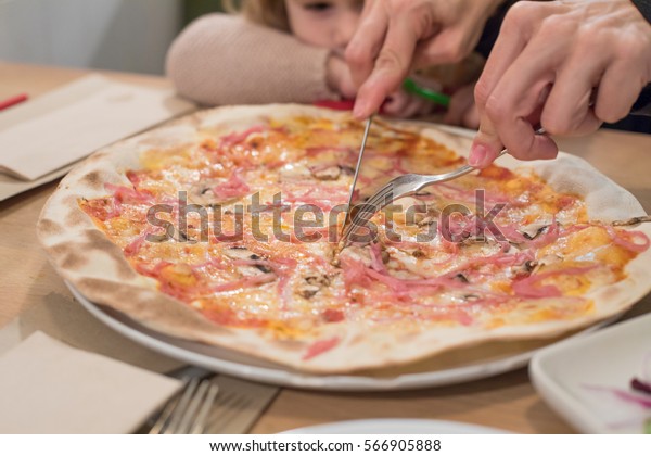 Three\
years old blonde child at restaurant table looking woman hands\
cutting ham and cheese pizza with fork and\
knife