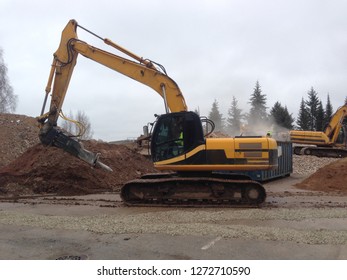 Three Yellow Construction Machines And  Men In High Visibility Work-wear Finishing Demolishing A Building With Piles Of Construction Waste, Gravel, Dirt  And Sand Around, Spruce Trees In The Back