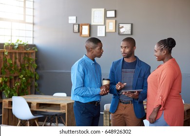 Three Young African Office Coworkers Smiling And Talking Together Over A Digital Tablet While Standing In A Large Modern Office