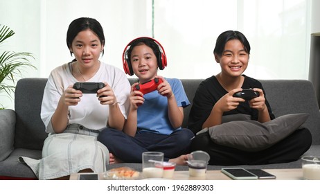 Three Young Asian Girls Playing Video Games Together On Sofa At Home.