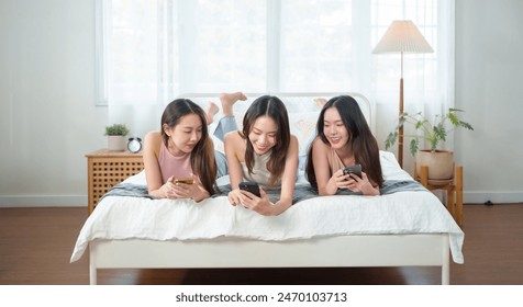 Three young Asian women lying on a bed using smartphones, displaying a relaxed and connected moment of friendship in a bright, cozy bedroom. - Powered by Shutterstock