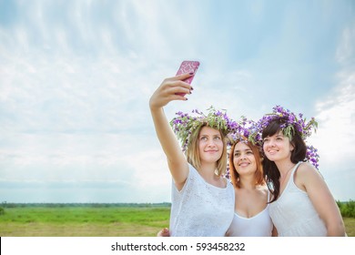 Three young beautiful girls in white dresses and wreaths of wild flowers doing selfie standing in a field. Summer in the village. Free space