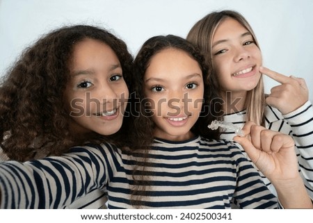 Three young beautiful multiracial kid girls make selfie holding an invisible braces aligner, recommending this new treatment. Dental healthcare concept.