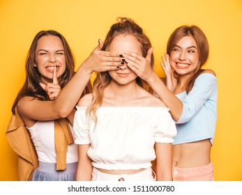 Three young beautiful smiling hipster girls in trendy summer clothes.Sexy carefree women posing near yellow wall in studio.Models surprising their friend.They cover her eyes and hugging from behind