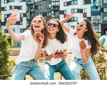 Three Young Beautiful Smiling Hipster Female In Trendy Summer  Clothes.Sexy Carefree Women Posing In The Street.Positive Models Taking Selfie In Sunglasses.Holding Juicy Burger And Eating Hamburger