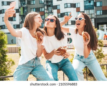 Three Young Beautiful Smiling Hipster Female In Trendy Summer  Clothes.Sexy Carefree Women Posing In The Street.Positive Models Taking Selfie In Sunglasses.Holding Juicy Burger And Eating Hamburger