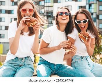 Three young beautiful smiling hipster female in trendy summer same clothes. Sexy carefree women posing in the street.Positive models having fun in sunglasses.Holding juicy burger and eating hamburger