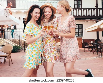 Three young beautiful smiling hipster female celebrating. Carefree women posing at summer terrace veranda cafe in the street.Positive models drinking champagne. Enjoying their vacation. Talking