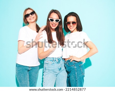 Three young beautiful smiling hipster female in trendy same summer white t-shirt and jeans clothes. carefree women posing near light blue wall in studio. Cheerful and positive models having fun