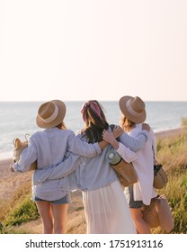 Three Young Beautiful Women Female Friends Going To Have Summer Picnic On A Beach At Sunset. View From Behind.