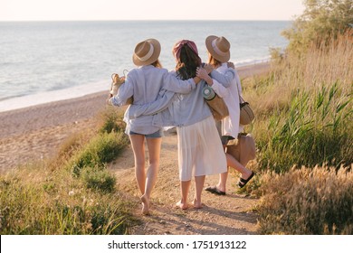 Three Young Beautiful Women Female Friends Going To Have Summer Picnic On A Beach At Sunset. View From Behind.