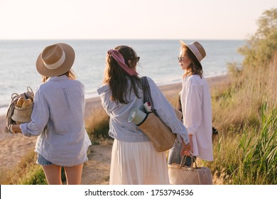 Three Young Beautiful Women Female Friends Going To Have Summer Picnic On A Beach At Sunset. View From Behind.