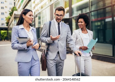 Three young business people talking to each other while walking outdoors with suitcase.