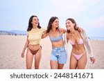 Three young Caucasian beautiful women friends embracing in swimwear posing cheerful on beach holidays. Group of gen z female hugging standing smiling on sunny summer time