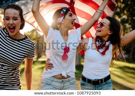 Three young female friends with American flag having fun in the park. Smiling group of women celebrating 4th of july outdoors.