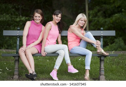 Three Young Female Friends Seated On A Bench, Outdoors In The Parc