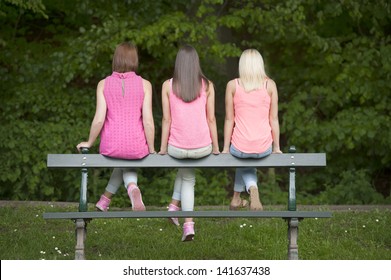 Three Young Female Friends Seated On A Bench, Outdoors In The Parc