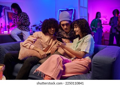 Three young friends in casualwear watching online videos in smartphone of cute brunette girl with curly hair while enjoying home party - Powered by Shutterstock