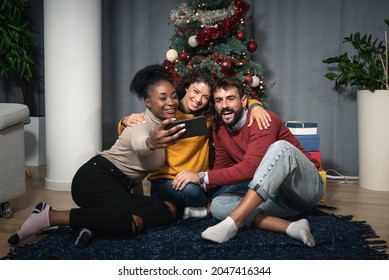 Three young friends celebrate Christmas and New Year's Eve together by joking and taking selfies with a smartphone in front of the Christmas tree waiting the midnight to exchange the gifts in boxes - Powered by Shutterstock