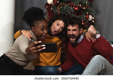 Three Young Friends Celebrate Christmas And New Year's Eve Together By Joking And Taking Selfies With A Smartphone Goofy And Making Funny Facial Expressions In Front Of The Christmas Tree