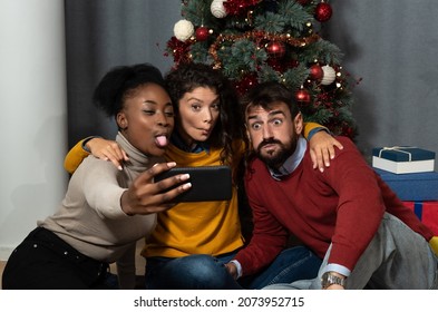 Three Young Friends Celebrate Christmas And New Year's Eve Together By Joking And Taking Selfies With A Smartphone Goofy And Making Funny Facial Expressions In Front Of The Christmas Tree