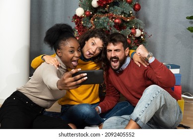 Three Young Friends Celebrate Christmas And New Year's Eve Together By Joking And Taking Selfies With A Smartphone Goofy And Making Funny Facial Expressions In Front Of The Christmas Tree