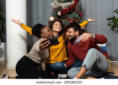 Three Young Friends Celebrate Christmas And New Year's Eve Together By Joking And Taking Selfies With A Smartphone Goofy And Making Funny Facial Expressions In Front Of The Christmas Tree