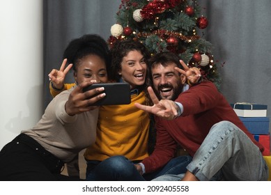 Three Young Friends Celebrate Christmas And New Year's Eve Together By Joking And Taking Selfies With A Smartphone Goofy And Making Funny Facial Expressions In Front Of The Christmas Tree