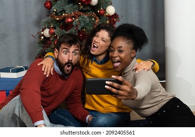 Three Young Friends Celebrate Christmas And New Year's Eve Together By Joking And Taking Selfies With A Smartphone Goofy And Making Funny Facial Expressions In Front Of The Christmas Tree