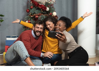 Three Young Friends Celebrate Christmas And New Year's Eve Together By Joking And Taking Selfies With A Smartphone Goofy And Making Funny Facial Expressions In Front Of The Christmas Tree