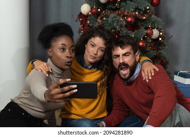 Three Young Friends Celebrate Christmas And New Year's Eve Holidays Together By Joking And Taking Selfies With A Smartphone Goofy And Making Funny Facial Expressions In Front Of The Christmas Tree. 