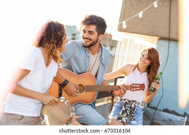 Three young friends having fun at rooftop party, playing guitar and singing. Focus on the man playing the guitar - Powered by Shutterstock