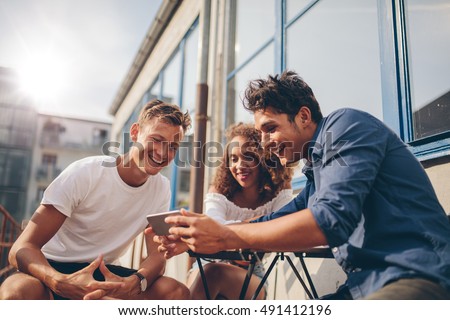 Three young friends sitting outdoors and looking at mobile phone. Group of people sitting at outdoor cafe and watching video on the smartphone.