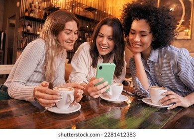 Three young friends using cell phone fun having drink at cafe shop. Smiling and happy girls looking at mobile while spending time together drinking coffee. Millennial women social networks.