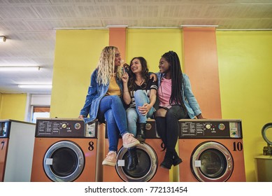 Three young girlfriends sitting together on washing machines in a laundromat laughing and blowing bubbles with chewing gum - Shutterstock ID 772131454